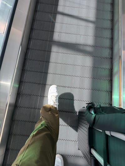 An upward-angle shot shows a person's legs and a suitcase on a moving escalator at Paris-Charles de Gaulle Airport in Le Mesnil-Amelot, France. The person is wearing white sneakers and olive green pants. They are positioned on the right side of the escalator with their right leg forward and their left leg behind. A dark grey suitcase with a green bag draped over it is to the person's right. Sunlight casts diagonal shadows across the escalator steps.