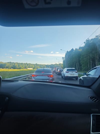 This media file shows a view from inside a car, looking forward at a traffic jam. The scene is set on a road with trees lining the right side and a grassy field on the left. The sky is clear blue with a few wispy clouds, suggesting it is daytime, likely late afternoon or early evening due to the soft lighting. The weather appears to be fair.
The primary subjects are the various cars in the traffic jam. We can see the back of a silver Kia sedan in the foreground, followed by a white Kia Sportage, and then several other vehicles including a van and darker sedans. The cars are stopped or moving very slowly. The activity is the progression of traffic, which is currently halted.
Notable details include the road infrastructure with a metal guardrail separating the lanes and streetlights visible in the distance. The interior of the car from which the photo is taken is partially visible, showing a dark dashboard. The license plate on the white Kia Sportage reads "P483KK 199", which appears to be a Russian license plate format. The overall mood conveyed is one of a typical traffic congestion, likely causing frustration for the drivers, although no specific emotions are discernible from the image itself. The setting is consistent with a highway or major road in Russia, as indicated by the provided location context.
