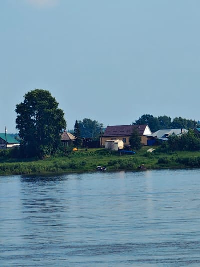 The image depicts a tranquil waterscape with a village nestled along the riverbank. The foreground is dominated by the rippling surface of a wide, blue river, reflecting the clear sky above. On the distant bank, a collection of houses with varying roof designs, some with prominent chimneys, are visible amidst lush green vegetation. A large, dense tree stands prominently on the left side of the bank, providing shade. Several small boats are moored along the shoreline, suggesting a connection to the river for transportation or recreation. The sky is a pale blue with subtle hints of clouds, indicating fair weather. The overall impression is one of a serene, rural setting in the city of Yeniseysk, Russia, a place where the river plays an integral role in the community's life.