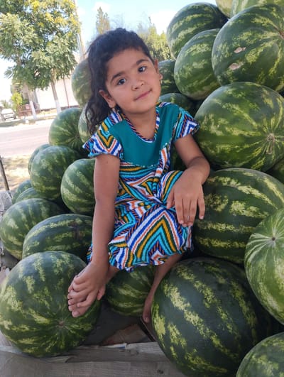 A young girl with dark hair sits cross-legged on a large pile of watermelons, her feet resting on one. She wears a teal dress with a colorful zigzag pattern. The watermelons are stacked high, with their green rinds and lighter stripes visible. The scene appears to be a market stall, with trees and a street visible in the background, suggesting an outdoor setting in a city. The lighting indicates it is daytime.