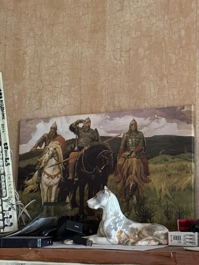 The scene depicts a section of a desk or shelf in what appears to be an indoor setting. A significant portion of the view is taken up by a textured, beige-toned wall or surface. A framed print or artwork is positioned toward the right. The artwork shows mounted figures, possibly soldiers or knights, in a field or landscape under a cloudy sky. The figures are dressed in historical armor and attire. To the right of the artwork is a small, white ceramic figurine of a horse, positioned among other objects. A USB hub or similar device, and other electronic cables are also visible. In the lower portion of the image, parts of a keyboard with handwritten markings are visible. The location of Ulyanovka, Russia, is not discernible from the image.