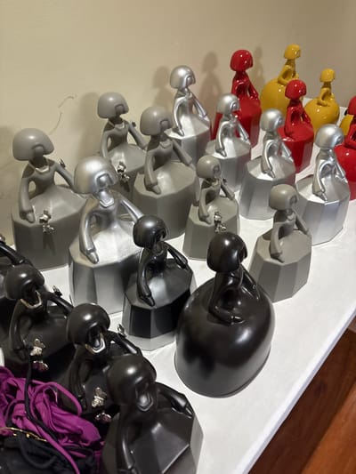 The image depicts a table covered in white cloth, in a room with beige walls. There are 14 decorative figurines displayed on the table. Most of the figurines are silvery-grey in color and feature female figures in traditional dresses. They are arranged in a row, with some of them stacked on top of each other. The remaining figurines are black and have a more modern design. The location of the image is Río Oro, Costa Rica, based on the coordinates provided (latitude: 9.945, longitude: -84.201). The figurines are possibly art pieces or souvenirs, as they are neatly arranged and on display in a room.
