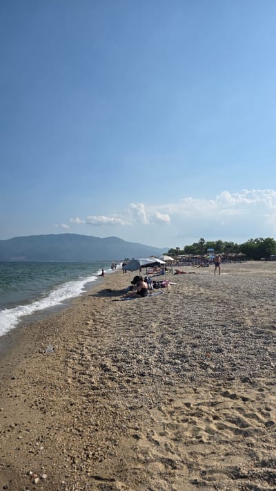 This is a vertical outdoor photograph depicting a busy beach scene on a clear, sunny day in Asprovalta, Greece. The upper portion of the image displays a vast expanse of light blue sky with scattered white cumulus clouds, primarily congregating on the right side. A prominent, elongated mountain range, appearing dark greyish-blue due to atmospheric haze, forms the distant background, stretching across the horizon.
The left side of the image features the Aegean Sea, characterized by its deep blue-green water and gentle waves breaking into white foam along the shoreline. Several individuals are visible swimming or wading in the water, dispersed along the coast.
The beach itself is composed of a mix of light brown sand and small, multi-toned pebbles, with the pebble concentration higher near the water's edge. Footprints are visible in the sand further from the sea. A small, white piece of plastic debris is located on the immediate shoreline in the lower-left quadrant.
Along the beach, numerous people are present. In the mid-ground, a person wearing a dark swimsuit is seated on the pebbly sand, partially shaded by a large, light grey beach umbrella. Adjacent to this individual are a baby stroller and several bags. Further to the right, a male wearing blue swim trunks is walking away from the camera, traversing the sandy area. Throughout the mid-ground and background, multiple groups of people are observed lounging on towels or sunbeds, many positioned under various beach umbrellas (grey, white, beige) that provide shade. A small, blue structure, consistent with a lifeguard stand, is visible in the mid-right background. A line of green trees and palm trees delineates the back edge of the beach, suggesting the transition to land. The scene is illuminated by bright, direct sunlight, indicating midday or early afternoon conditions.