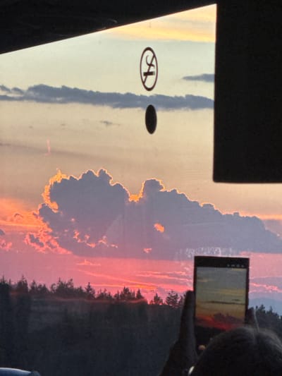The image shows a view from inside a vehicle looking out a window at a sunset over a landscape. A no-smoking sign is visible on the window. A person's hands are holding a smartphone, taking a picture of the sunset, which is reflected on the phone's screen. The sunset features vibrant pink and orange hues in the clouds and sky, with a silhouette of trees and hills in the foreground. The specific location in Baliovtsi, Bulgaria, is inferred from the context provided, but not directly visible in the image itself.