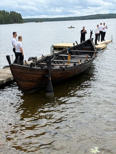 A wooden rowboat is docked at a wooden pier on a lake in Aneby, Sweden. The boat is dark brown and appears to be old, with rivets visible along its hull. An oar is partially submerged in the water next to the boat. Several men are standing on the pier, some are holding phones, and appear to be looking out at the water. The sky is overcast, and the surrounding area is forested.