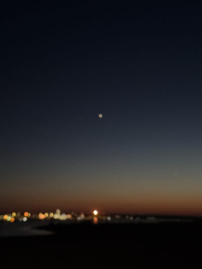 This image captures a nighttime scene with a dark, gradient sky transitioning from deep blue to a hazy orange near the horizon. In the upper left quadrant of the sky, a small, bright celestial body is visible. The right side of the image features a blurred vertical line of bokeh lights, suggesting the presence of a city or man-made structures at a distance. The lights appear in various colors, primarily yellow, white, and orange, and are out of focus, creating a dreamlike effect. The overall impression is one of twilight or early night, possibly with some atmospheric haze. No people or specific objects are clearly discernible beyond the celestial body and the bokeh lights. The setting appears to be an outdoor environment, likely overlooking a distant, illuminated area.