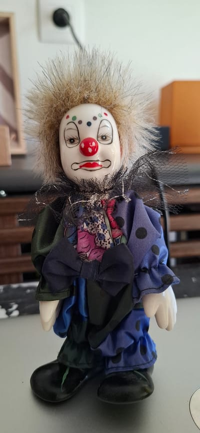 The media file shows a close-up, rotated view of a porcelain doll designed as a clown. The doll is dressed in a layered, colorful costume featuring a blue fabric with black polka dots, patterned fabric with floral designs in pink and blue, and dark green fabric. The clown has a white porcelain face with painted features, including wide eyes, red cheeks, a red circular nose, and painted red tears or lines below the eyes. It has a large, fluffy, light brown wig and wears a black net, possibly part of its costume, around its head and neck. The doll is positioned as if lying down, with its head to the left and its feet to the right. The background is blurred, showing hints of wooden furniture and a light-colored wall with a white electrical outlet. The lighting appears to be artificial and indoor. There is no discernible text in the image. The overall impression is of a decorative item displayed indoors, possibly on a shelf or table. The doll itself does not appear to be engaged in any activity. The setting is suggestive of a domestic or retail environment.