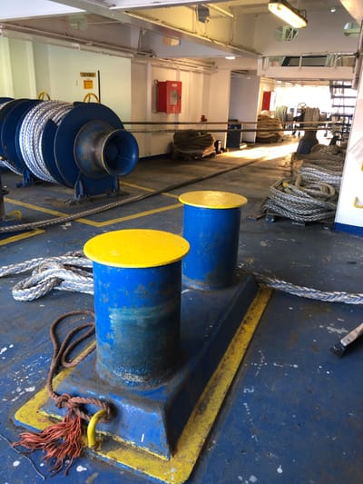 The image depicts the internal deck of a ship, located in La Spezia, Italy, featuring various maritime mooring equipment. In the foreground, two blue cylindrical bollards with bright yellow caps are bolted to the worn blue deck, outlined by yellow painted lines. A frayed brown rope with red ends is knotted around a yellow hook on the base of the left bollard. A thick white and blue braided rope lies coiled on the deck and extends across the foreground, alongside a small paintbrush on the right. The blue deck surface exhibits wear, including white splatters and scuff marks.
In the midground, a large blue winch spooling white braided rope is positioned on the left. An integrated blue, trumpet-shaped fairlead is part of the winch assembly, from which a steel chain extends across the deck. A red fire extinguisher cabinet is mounted on a white bulkhead. Further coils of thick rope are stacked on wooden pallets to the right. Sunlight illuminates sections of the deck from an opening further aft. A yellow sign marked "OXYGEN" is affixed to the bulkhead above the winch. Overhead, white bulkheads, structural beams, and fluorescent light fixtures are visible. No individuals are present in the scene.