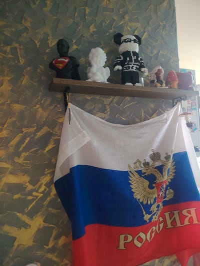 A Russian flag is displayed prominently in front of a textured wall. On a wooden shelf above the flag, several decorative figurines are visible. From left to right, these include a bust of Superman, a white cherub figurine, a black and white panda figurine, a figurine of a woman with blonde hair, and a mushroom-shaped decoration. The overall impression is of a decorated interior space with patriotic and decorative elements.