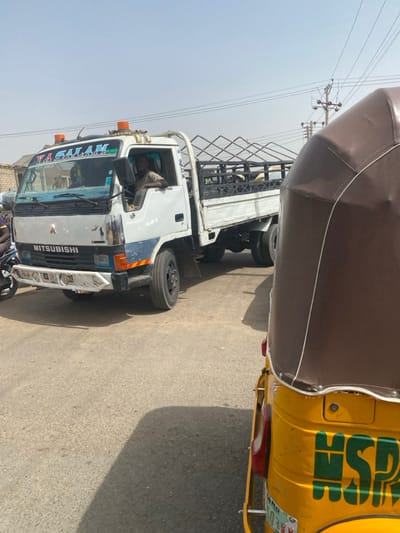 Located in Rijiyar Lemo, Nigeria, the image captures a street scene under clear daylight. A white Mitsubishi flatbed truck is prominent on the left, displaying "YA SALAM" on its windshield visor and two orange rotating beacons on the cab roof. The truck's front bumper is silver and shows visible wear, with blue and yellow/orange accents on the lower front body. A male driver, wearing a light-colored shirt, is seated in the driver's position, looking forward. Another individual is partially visible in the passenger seat. The truck's white flatbed is equipped with a black metal cage structure. In the immediate foreground on the right, the rear-right portion of a yellow trishaw, topped with a brown canopy, partially obstructs the view. Green "HSP" lettering is affixed to the trishaw's yellow body. The ground consists of an asphalt road surface with shadows cast upon it. Utility poles with power lines are visible in the background on the right, and distant buildings are partially seen on the left. A motorcycle is partially visible behind the truck on the far left.