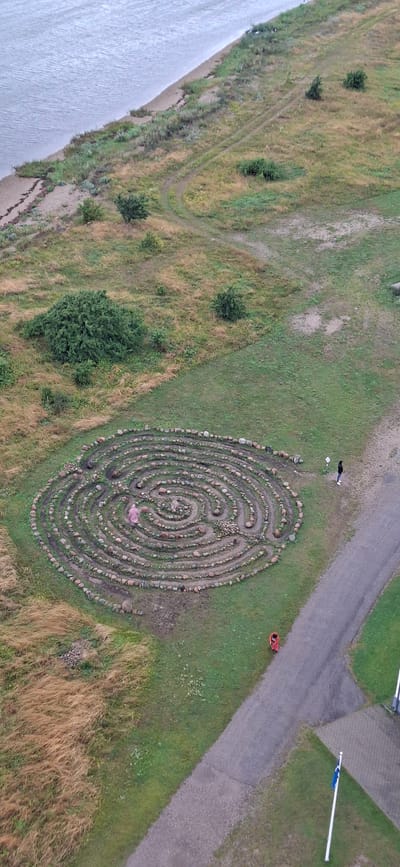 In the city of Sääre, Estonia, an aerial view captures a circular stone labyrinth situated on a grassy hill. The labyrinth's intricate design is formed by a winding path of stones, encircling a central area. A person dressed in pink is visible within the labyrinth, walking along the path. To the right of the labyrinth, a paved path leads away, and two other individuals are present on this path. One person, wearing a dark jacket and white shoes, stands near the labyrinth, while another, dressed in red, is further down the path. The surrounding environment is a mix of green grass, dry, tall grasses, and sparse bushes. In the distance, a body of water borders the landscape, with a sandy shoreline. A flagpole with a blue and white flag is positioned in the lower right corner of the image, adjacent to a paved area.