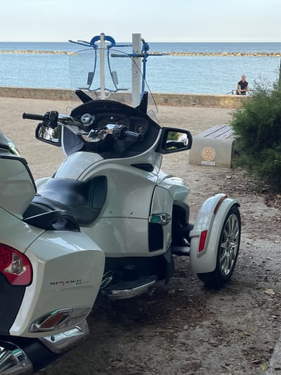 The image shows a parked Can-Am Spyder RT, a three-wheeled motorcycle, near the beach in Germasogeia, Cyprus. The bike is white and faces toward the coastline. In the background, across a narrow strip of beach, is the sea, and a breakwater extending into the water. A person is visible sitting on a bench near some vegetation, positioned behind the motorcycle. A concrete bench or structure with a Rotary International emblem is also near the vegetation. The ground around the motorcycle is sandy and covered with small leaves or debris. The image is oriented vertically.