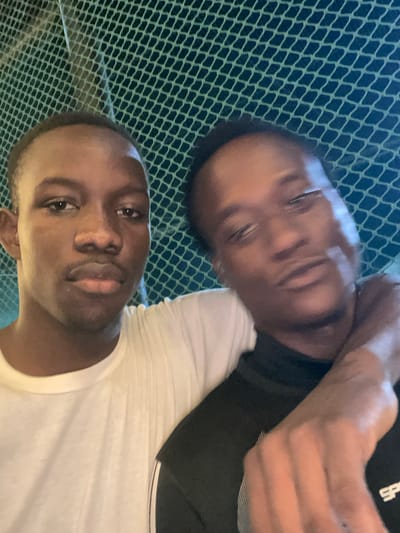 The media file shows a close-up, tilted selfie of two young Black men lying down. The man in the foreground, wearing a white t-shirt, is looking directly at the camera with a neutral expression. His face is in focus, highlighting his dark eyes and lips. Behind him, the upper body of another man is visible, also looking towards the camera. He is wearing a black t-shirt with white text on the sleeve, which appears to be a logo or brand name. His arm is resting on what seems to be the other man's shoulder or head, and his facial features are slightly blurred.

The background consists of a green netting with a diamond pattern, suggesting an outdoor setting like a sports court or a fence. The lighting appears to be artificial or dim, possibly indicating it's evening or indoors. The overall mood is casual and intimate, typical of a selfie taken between friends. The location context provided is Malete, Nigeria. There is no indication of the weather or specific time of day beyond the general lighting conditions. The visible text on the black t-shirt is partially obscured but contains white lettering.