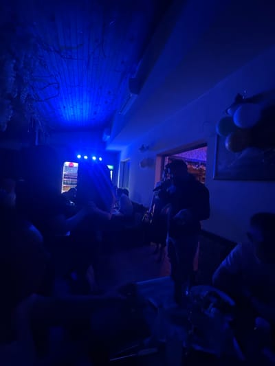 The image shows an indoor scene with a dark ambiance illuminated by blue lighting. A brick wall is visible on the left. In the background, there are a few people gathered in what appears to be a room. There are balloons hanging from the ceiling. Based on the location, the image is likely taken in Panagyurski kolonii, Bulgaria.