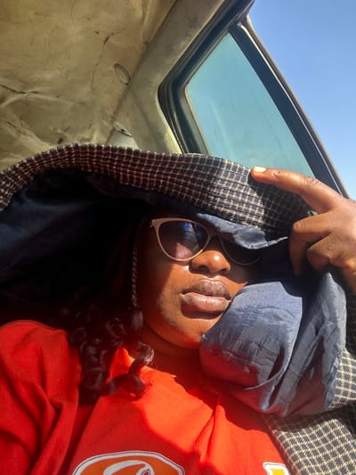 An adult individual with dark skin, wearing light-framed sunglasses and an orange top, is depicted in a close-up inside a vehicle in Rimau, Nigeria. The person is actively using a brown and black houndstooth pattern jacket, lined with dark blue fabric, to shade their face from direct, intense sunlight entering through a window. A finger is positioned on the upper edge of the jacket, maintaining its shading position. The vehicle's interior shows a worn, light-colored headliner above, and a clear, bright blue sky is visible through the adjacent window, indicating a sunny environment.
