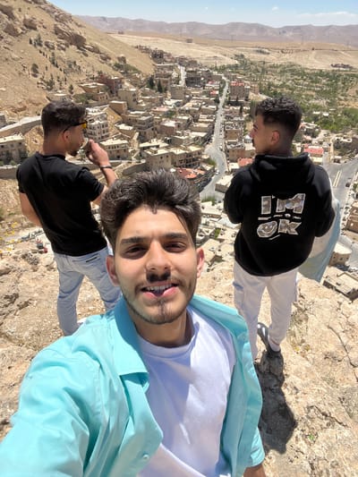 Three young men are on a precipice overlooking a town in Ma'loula, Syria. The man closest to the camera is wearing a white t-shirt, a light blue button-down shirt, and light blue jeans. He is facing the camera, partially smiling. Behind him, to the left, is a man wearing a black t-shirt and light blue jeans. A third man is facing away from the camera, wearing a black hoodie with a design on the back and light-colored pants. The town below is composed of closely packed buildings and roads. The sky is clear and sunny. The terrain is rocky and arid.
