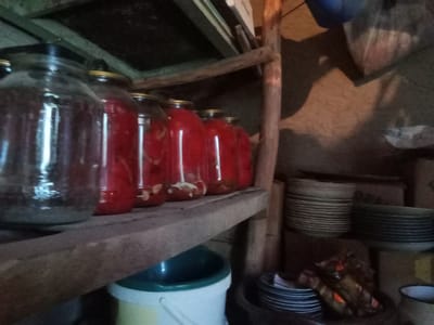This is a medium shot of a dimly lit, rustic pantry or storage area, likely in Karaganda, Uzbekistan. On a rough wooden shelf in the foreground, several large glass jars are filled with a red liquid and what appear to be preserved fruits or vegetables. To the right of the jars, stacked on a lower shelf, are several stacks of plates. The walls are rough and textured, and a wooden support beam is visible on the right side of the image. The overall impression is one of a functional, perhaps traditional, food storage space.
