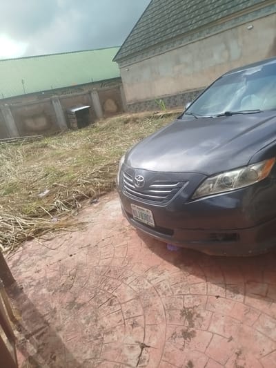 A dark gray Toyota Camry is parked on a patterned concrete driveway in Isiehor, Nigeria. The car's front end is visible, including the grille, headlights, and license plate which reads "ABUJA KJU-830G". The car is positioned to the right of the frame. To the left of the car, dry, overgrown grass and some scattered debris are present. In the background, a stone wall with metal fencing runs horizontally. Beyond the wall, a building with a green, corrugated metal roof is visible, as is another building with a gray tiled roof. The sky above is overcast.