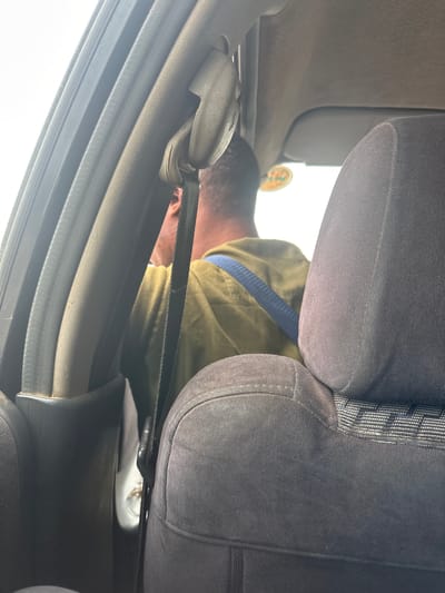 The image shows a man inside a vehicle, possibly a car, from a low angle looking upwards. He is wearing a green jacket with a blue stripe across it. The man is leaning forward, with his head obscured by the car's seat. The interior of the car appears to be dark grey. A seatbelt is visible, stretched across the scene, and a part of the car's door frame is also visible on the left side. The lighting suggests it is daytime, with bright light coming from outside the vehicle, indicating it might be sunny. There is a circular sticker visible on the exterior of the car, near the man's head, but the text is too blurry to be legible. The setting is likely a street or a parking area in Crescent, Nigeria. The overall mood is casual, with the man possibly searching for something or attending to something within the car.