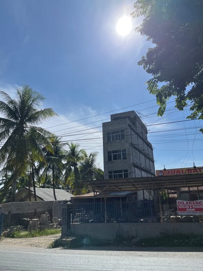 The image depicts an outdoor scene in what appears to be a semi-developed area in Bukitrata, Indonesia. The foreground shows a paved surface, possibly a road, with a grassy verge. Behind this, a fence encloses an area with various construction materials, including stacks of concrete blocks and what looks like decorative stone or concrete elements. Adjacent to this is a low-lying structure with a corrugated metal roof, and beyond that, several palm trees are visible.

Dominating the mid-ground is a multi-story building under construction. Its concrete facade is unfinished, with exposed rebar protruding from the upper sections. Windows are installed in some floors, and the building appears to be several stories high. To the right of this building, a sign with red lettering reads "DILARANG JUALAN & PARKIR DI SINI" and "JANGAN BUANG SAMPAH DI SINI," indicating a prohibition against selling, parking, and dumping trash. Another sign above this reads "JUAL BATU" which translates to "selling stone."

In the background, more palm trees are scattered, and the sky is bright blue with a few wispy clouds. The sun is high in the sky, casting strong light and creating some lens flare in the upper right quadrant of the image. Overhead, multiple power lines crisscross the scene, supported by poles that are not fully visible. The overall impression is of a tropical environment in a state of ongoing development.