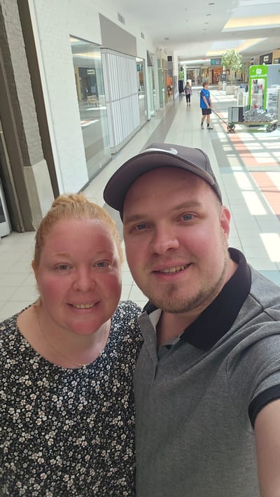 In this selfie taken inside a mall, a man and a woman pose for the camera. The man, wearing a grey polo shirt and a baseball cap, stands on the right, while the woman, in a black floral dress, is on the left. Behind them, the mall corridor stretches into the distance, with shops on either side. The flooring is tiled in a pattern of light and dark rectangles, with a slight sheen. Sunlight streams in from the ceiling, creating distinct shadows. In the background, a young man in a blue vest pushes a cleaning cart, and a few other shoppers can be seen walking. The mall appears to be the Laurier Quebec, located in Victoriaville, Canada.