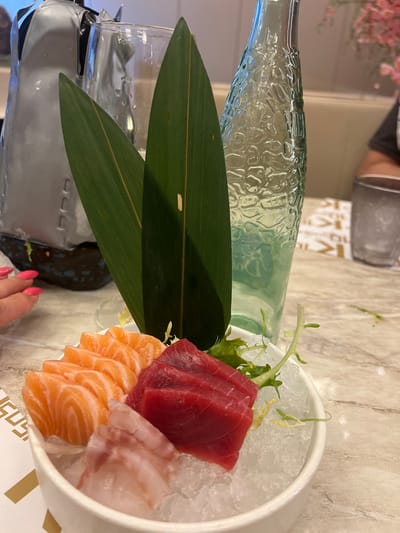 The image presents a close-up view of a sushi platter. The platter, a white bowl filled with crushed ice, contains an assortment of sliced raw fish including tuna (red), salmon (orange), and possibly sea bass (white). Two large green leaves are placed on the side of the bowl as garnish. A glass bottle filled with a clear liquid, likely water, is seen in the background to the left of the bowl. The scene appears to be indoors, on a table, with a marble-patterned surface. A portion of a person's hand with pink nail polish is visible in the lower right corner. A small section of what seems to be a white and gold patterned table cloth is visible. The location in Torreforta, Spain is not apparent from this image.