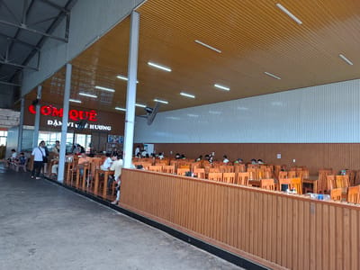 This image depicts the interior of a large, open-air restaurant, likely a food court or a family-style eatery, located in Tân Phú 2, Vietnam. The restaurant features a prominent sign with red lettering reading "CƠM QUÊ" and smaller text below it, indicating a focus on traditional or home-style rice dishes. The seating area is extensive, with numerous wooden tables and chairs arranged in rows. A light brown wooden wall with vertical paneling forms a partition in the foreground, separating the dining area from the walkway. The ceiling is a warm, honey-colored material with a ribbed texture, illuminated by long, rectangular fluorescent lights. The background shows a more enclosed section of the restaurant with patrons seated at tables and interacting. A fan is visible mounted on a pole near the ceiling, suggesting efforts to manage the environment's temperature. The overall atmosphere appears to be casual and busy, with many people dining.