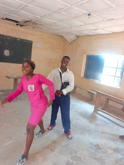 The scene depicts two young individuals within a classroom in Mada, Nigeria. A female, positioned in the foreground and to the left, is captured in motion, leaning forward with her arms slightly bent and legs extended, smiling broadly. She wears a pink long-sleeved dress with a white patch on the chest, a patterned headscarf, and grey and white patterned clogs. Behind and to her right stands a male, smiling broadly towards the camera. He wears a white long-sleeved collared shirt, dark blue trousers, red and black slides, and a black messenger bag slung across his chest.

The classroom features light-colored walls extensively covered with white chalk inscriptions, including names, dates such as "12-2-2024," and phrases like "M. SALAH," "EL CAMPEON," "LIVE TO REMEMBER," and "ISTIFANUS D.D.O." A dark blackboard with visible chalk markings is mounted on the left wall, behind the female. To the left, a wooden table is partially visible. Along the right wall, several basic wooden benches are arranged, suggesting seating. A dark-framed window on the right wall admits bright, natural light, creating a glare. The ceiling is constructed from white tiles, with several panels damaged or missing, and a bare light socket hangs from the center. The concrete floor exhibits wear and discoloration.