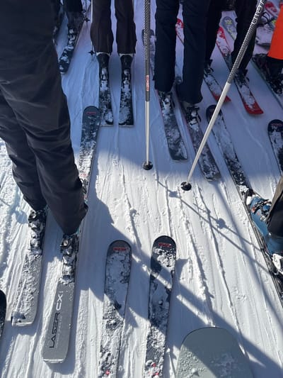 The image displays a ground-level perspective of multiple individuals engaged in skiing or snowboarding activities on a groomed snow surface in Zürs, Austria. Numerous pairs of skis, including a pair labeled "LASER CX" in the foreground, and at least one snowboard are visible. The skis are varied in color, with some featuring red and black details, and are partially dusted with fresh snow. Ski boots, such as a prominent blue pair on the right, are secured into bindings. Several ski poles, including one with a carbon fiber pattern and another white/silver pole, are planted upright in the snow. Long, defined shadows extend from the poles and individuals towards the lower right of the frame, indicating direct sunlight and a clear outdoor environment. The individuals are primarily visible from the waist down, wearing dark-colored ski pants. The arrangement of skis suggests a waiting area or a queue.
