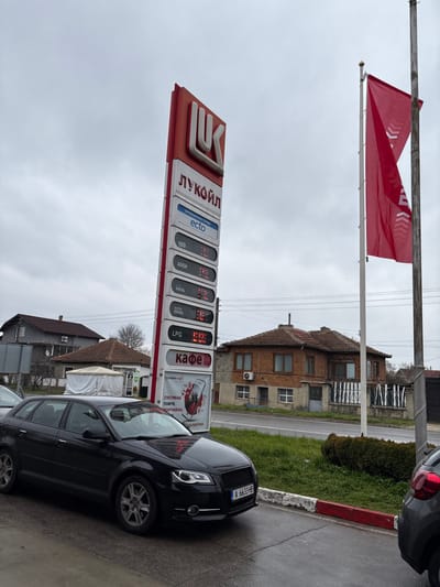 The image displays a Lukoil gas station in Staro Oriahovo, Bulgaria, under an overcast sky. A vertical red and white Lukoil price pylon stands centrally, indicating fuel types and prices: "Ecto" (---), "100" (3.19), "A95H" (3.09), "Diesel" (3.10), "Super Diesel" (3.12), and "LPG" (1.25). Below the fuel listings, a panel is labeled "Кафе" (Cafe), followed by a digital advertisement at the bottom. To the right of the pylon, a red flag bearing white Lukoil branding is affixed to a pole.
In the foreground, a black Audi A3 sedan, bearing Bulgarian license plate A 6633 HB, is parked on wet asphalt, oriented towards the right. The front section of a second dark-colored vehicle is visible to the immediate right of the Audi. The ground in the foreground includes a curb with distinct red and white paint markings and small patches of green grass.
In the background, across a visible paved road, multiple residential buildings are present. These structures feature reddish-brown tiled roofs and facades of brick or stucco. One house displays visible windows and an outdoor air conditioning unit. A white, temporary tent-like structure is also discernible among the background buildings. No individuals are visible within the scene.