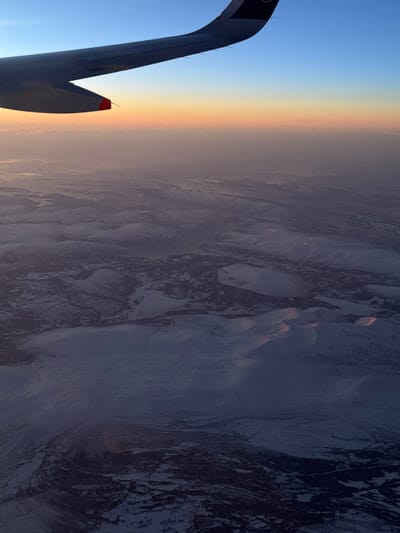 An airplane wing, with a visible black emblem, slices into a blue sky transitioning to a hazy, golden-toned horizon. Beyond, a rugged landscape of snow-dusted mountains and deep valleys stretches out. The scene is captured from a high vantage point, possibly mid-flight, offering a vast view of the Earth's surface. The texture of the terrain contrasts with the smooth, manufactured surface of the aircraft wing.
