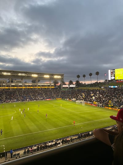 The image shows a soccer game at Dignity Health Sports Park in Carson, California, USA. The stadium is filled with spectators on both sides of the field. The soccer game is in progress, and the players are visible on the green field. The sky is overcast with grey clouds, though there are hints of sunset beyond the palm trees. Advertisements surround the stadium, including "Modelo," "Coca-Cola", and "Dignity."