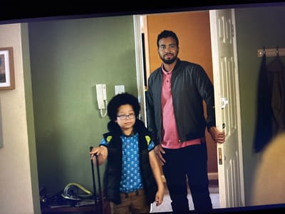 An indoor scene captures a man and a child standing in a doorway or entryway. The man, on the right, is light-skinned with dark, short curly hair and a beard. He wears a dark grey bomber jacket over a pink polo shirt and dark trousers, and holds a white, paneled door ajar with his right hand. He is looking towards the viewer with a slight smile.
To his left stands a young boy with dark, curly hair and glasses, also looking towards the viewer with a neutral expression. He wears a blue polo shirt with a white pattern, a dark puffer vest with yellow accents on the shoulders, and light brown trousers. He holds the extendable handle of a small piece of luggage or a bag on wheels, which is partially visible at the bottom left.
The setting appears to be a home's entrance hall. To the left, a muted green wall features a white intercom unit. Further left, a lighter beige-colored wall has a framed picture. Behind the man, through the open door, an orange-brown wall is visible. To the right of the doorway, against another green wall, there is a wooden coat rack with a dark jacket or coat hanging on it. On a surface near the child's feet, various items are scattered, including what looks like a coiled yellow hose or cable and some papers. No visible text is present in the image. The lighting is general indoor illumination, suggesting an ordinary moment within a residence in Dolna banya, Bulgaria.