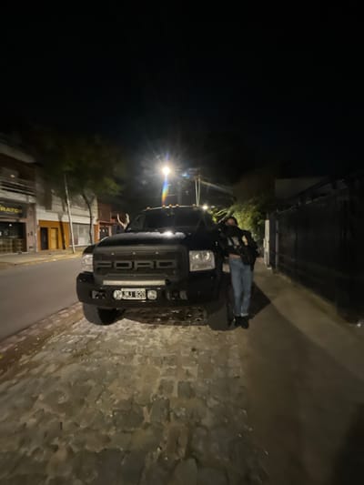 At night, a black Ford truck is parked on a street with buildings on the left side and a wall on the right. A person is sitting on the hood of the truck. The license plate of the truck reads "NMJ 820." The scene appears to be in a residential area. Based on the street signs and architectural style, the location is likely in the city of Olivos, Argentina.