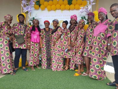 The image shows a group of ten people, all wearing brightly colored clothing, standing in front of a wall with the inscription "First Baptist Church Teenage Department". They are standing on a green lawn with a wall to the right, decorated with yellow balloons and flowers. The people are positioned in a semi-circle, with the exception of one man wearing a black shirt who stands to the left of the group. The group is likely from a church or a school, and they appear to be posing for a photo. The coordinates provided correspond to a location in Yenagoa, Nigeria. The scene is likely taking place in front of the First Baptist Church in Yenagoa.