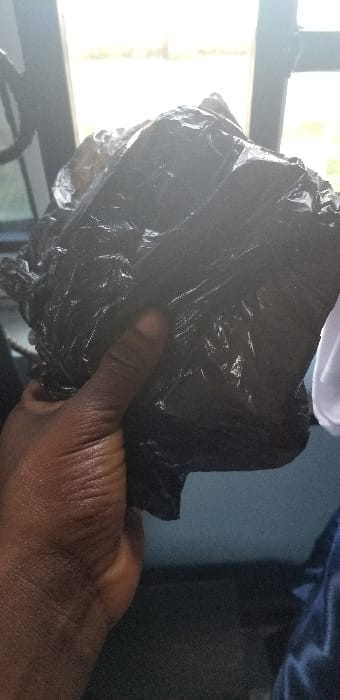A dark-skinned human hand is shown grasping a crinkled black plastic bag. The thumb and fingers are visible, securing the bag on the left side of the frame. The black plastic bag contains an irregularly shaped, lumpy mass, obscuring its contents. The bag's surface is reflective. The background is out of focus, featuring an overexposed bright rectangular area in the upper portion, consistent with a window or light source. A dark vertical structural element is present to the left of this bright area. Indistinct light blue and grey tones are visible in the lower right background. The scene is located in Ado-Ekiti, Nigeria.