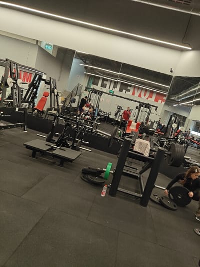 This image displays the interior of a modern fitness center or gym located in Nahariya, Israel. The facility features dark rubberized floor tiling, light gray walls, and a prominent mirrored wall reflecting the gym's layout, overhead linear LED lighting fixtures, and a large red text graphic that reads "LOVE LIFE" along with some reflected Hebrew text, possibly an exit sign.

Numerous pieces of exercise equipment are visible, including multiple weight machines, barbells, various black and green weight plates, and benches, one featuring red upholstery. In the foreground on the right, a female individual with long brown hair, dressed in dark athletic wear, is crouched and handling a black weight plate, either placing or removing it from a barbell setup. A clear plastic water bottle with a red cap is positioned on the floor nearby. In the midground and reflections, approximately six to eight other individuals are dispersed throughout the gym, engaged in or preparing for exercise at different stations. A white towel is draped over a piece of equipment in the mid-right.