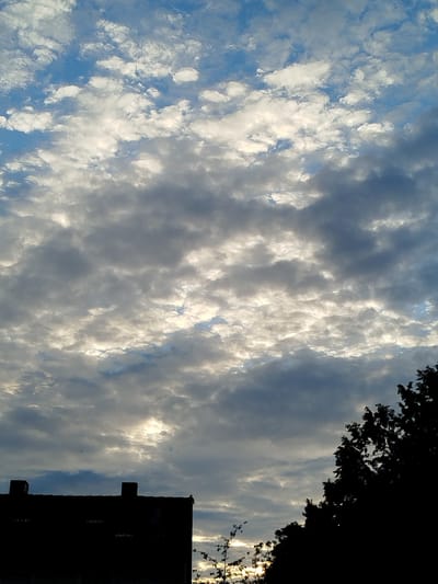 The image shows a cloudy sky above a silhouetted building and trees. The sky is a mix of blue and gray clouds, with patches of sunlight breaking through. The building, which appears to be a residential structure with chimneys, is in the lower left of the frame, completely dark. To the right, the silhouettes of trees are visible, their branches extending upwards. The scene suggests an outdoor view, possibly from a window or an open area in Kitzingen, Germany. There are no people or animals visible. The overall impression is one of a partly cloudy day, with the sun low in the sky, casting shadows and creating a dramatic contrast between the bright sky and the dark foreground elements.