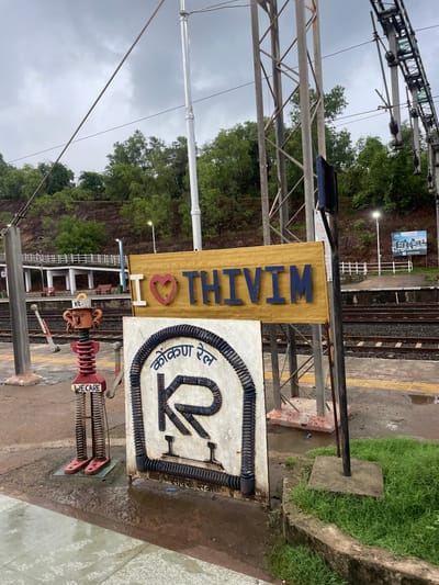 This image shows a sign at a train station in Sircaim, India. The sign is golden yellow with the word "THIVIM" written in blue letters. Below "THIVIM" is a red heart. To the right of the "THIVIM" sign is a circular logo with blue lettering that appears to be in an Indian language, along with a large "RK" symbol. The sign is mounted on a pole. There are also some decorative elements at the base of the sign, including a red figure with springs for limbs that has the word "WE CARE" written on its chest.

The setting is an outdoor train station platform. There are train tracks visible behind the sign, as well as a covered waiting area with benches in the background. Green trees and a rocky embankment are visible behind the station structures. Overhead, there are electrical wires and support structures for the train lines. The sky is overcast, suggesting a cloudy day. The ground of the platform is made of concrete and appears wet in places, indicating recent rain. The image is taken from a low angle, looking up towards the signs and the overhead wires. The overall mood is one of a functional, yet somewhat decorative, public space.