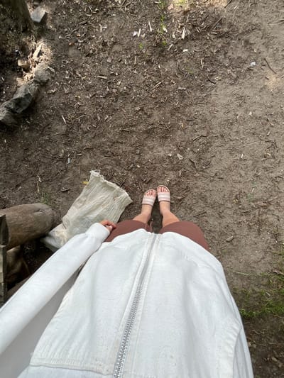 The media file is a top-down, eye-level shot of a person's legs and feet, standing on a dirt ground littered with small sticks and debris. The person is wearing pink and white striped slides and a brown skirt, with a white zippered hoodie or jacket over their legs. Part of a rough, off-white fabric, possibly a bag or blanket, is visible on the ground near their feet, along with a section of a rough wooden log.
The scene appears to be outdoors, in a natural or semi-natural environment given the dirt ground and scattered organic matter. The lighting suggests it is daytime, with dappled light indicating sunlight filtering through trees or foliage. No specific emotions are conveyed. There is no visible text in the image. The location context provided is Вяззеўскі сельскі Савет, Belarus.