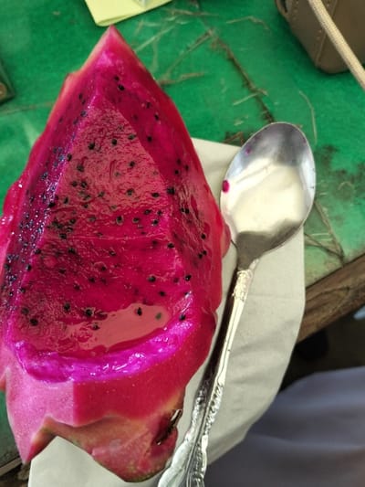 A vibrant, triangular-cut red dragon fruit, speckled with tiny black seeds, rests on a white napkin beside a silver spoon. The fruit's juicy interior glistens, with a slight pool of liquid at its base. The napkin sits on a weathered wooden surface, partially covered by a green surface and hints of a beige object in the upper right corner.