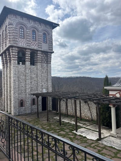 This image captures an outdoor scene in Nepraznentsi, Bulgaria, featuring a stone bell tower, a pergola, and distant hills under a cloudy sky.
In the foreground, a black wrought iron railing with intricate scrollwork runs horizontally across the lower third of the frame.
The dominant structure is a tall, multi-story stone bell tower, constructed from irregular light-colored stone blocks and horizontal bands of red brick. Its lower section includes two small arched windows with red and white striped surrounds. The mid-section features two large, open arched bays housing dark bells. The upper section displays three arched windows with dark frames, set against decorative red and white brickwork patterns, including 'X' shapes and diamond motifs. The tower is capped with a dark, slightly overhanging roof.
Extending from the bell tower to the right is a wooden pergola structure, composed of dark wooden posts supporting a lattice of bare, twisted vines. The ground beneath the pergola is an uneven surface of large, irregularly shaped flagstones interspersed with patches of green grass and dirt. A low stone wall, mirroring the construction style of the bell tower, runs along the rear of the pergola.
To the far right, a portion of another building is visible, featuring a white architectural column with a red-tiled base and a section of a dark, curved or domed roof. Behind this structure, a tall, slender coniferous tree stands prominently.
The background consists of a sprawling expanse of hills or mountains covered in dense, bare deciduous trees, indicating a winter or early spring season. The sky is largely overcast with prominent grey and white clouds, with glimpses of lighter blue sky in the upper right. No individuals or specific actions are depicted within the scene.