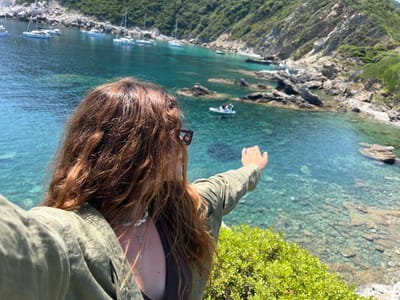 A woman with long, reddish-brown hair is standing on a cliff overlooking a bay filled with crystal-clear turquoise water. She is wearing a green jacket and sunglasses, and her right arm is outstretched, as if pointing or gesturing. In the bay, several white sailboats and a small inflatable boat are anchored. The coastline is rocky and covered with lush green vegetation. The sun is shining, casting a bright light on the scene. The image appears to be taken at a scenic overlook near Loutraki, Greece, given the characteristic clear waters and mountainous coastline often found in the region.