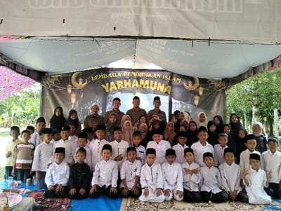 The image presents approximately 48 individuals, predominantly children, gathered for a group photograph under a temporary canopy structure in Jambo Dalam, Indonesia. The setting is semi-outdoor, with lush green trees and foliage visible in the background beyond the canopy.
Overhead, a white fabric canopy is suspended, and a prominent black banner stretches across the rear, featuring the text "LEMBAGA PENDIDIKAN ISLAM YARHAMUNA" in yellow lettering, accompanied by decorative motifs of a crescent moon, stars, and hanging lanterns. A segment of a pink patterned fabric panel is visible on the canopy's left side.
The group comprises five adult males standing in the back row, several adult or older teenage females, and a larger number of children of varying ages. The adult males wear a mix of patterned shirts, some resembling uniforms, with dark or white pecis (caps). The females are mostly attired in hijabs and modest clothing. The children, ranging from elementary to early adolescent age, are arranged in multiple rows. Many boys in the front rows are kneeling or squatting, typically wearing white long-sleeved tunics, dark pants, and dark pecis. Girls are interspersed throughout the group, also in modest attire with hijabs. All individuals are oriented towards the camera. One boy in the front row, on the far right, holds up a "V" sign with both hands.
The ground beneath the individuals is covered with blue and patterned rugs or mats. In the left foreground, a low surface displays several plastic cups containing liquids and yellow beverage cans.