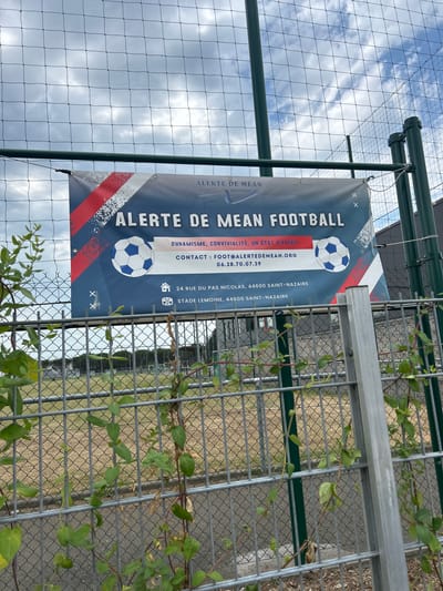 The image shows a banner on a chain-link fence in front of a grassy field. The banner reads "ALERTE DE MEAN FOOTBALL" and provides contact information, including an email address "FOOT@ALERTEDEMEAN.ORG" and a phone number "06.28.70.07.39". Below this, it lists an address "24 RUE DU PAS NICOLAS, 44600 SAINT-NAZAIRE" and then "STADE LEMOINE, 44600 SAINT-NAZAIRE". The banner also features images of soccer balls and the French flag colors. The environment appears to be an outdoor sports facility, possibly a soccer field. The sky is cloudy. There are no people visible in the image.