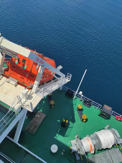 The image provides an overhead, slightly angled view of the deck of a large vessel, likely a ship or ferry, set against the calm, deep blue waters off Kunak, Malaysia. The vessel's main deck is painted a vibrant green, with various white structures and equipment.

A prominent orange enclosed lifeboat is situated on the mid-left side, mounted on a white davit system ready for deployment. The davit arm is visibly marked with "SWL 59KN", indicating its Safe Working Load in kilonewtons. The lifeboat features small windows and standard green exit signs.

Towards the forward section of the deck, various marine equipment is arranged. A large grey mooring winch or capstan holds a substantial coil of thick white rope, secured with an orange band. Nearby, a black bollard with yellow and black hazard stripes is visible, along with another coil of white rope on a similar mechanism. A red and white lifebuoy hangs from a white railing. A long, white antenna-like mast with a red cap extends outwards from the deck over the water. Other deck features include metallic grated platforms, a wooden pallet, and various black boxes and small white circular devices. One black piece of equipment near the bow is marked "SWL 25 TON 20C".

The strong, distinct shadows cast by the ship's structures suggest bright sunlight, indicating a time of day likely around midday or early afternoon. The water exhibits gentle ripples, signifying calm weather conditions. No individuals are visible in the frame, presenting a static snapshot of the vessel's operational deck.