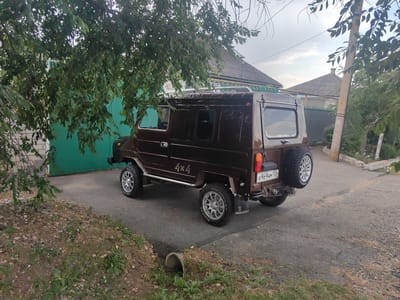 A dark brown, off-road vehicle with "4x4" markings is parked on a paved area beside a residential property. The vehicle has a spare tire mounted on the rear and a license plate that reads "A969HM 126 RUS". A green garage door is visible behind the vehicle, and a house with a gray roof is situated further back. Trees with green leaves partially obscure the scene. The ground is a mix of asphalt, gravel, and sparse vegetation. A drainage pipe is visible in the foreground. The sky is overcast.