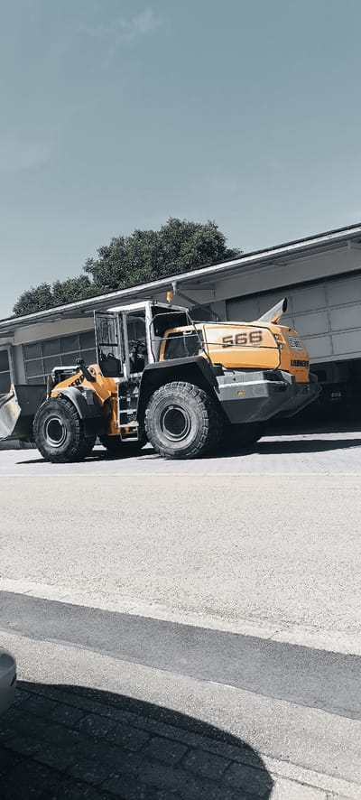 A yellow and gray Liebherr wheel loader with the number "586" on its side is parked on a paved surface in front of a building with a large garage door. The building appears to be a commercial or industrial structure with a white facade and a dark roof. Trees with green foliage are visible behind the building, and a clear blue sky with a few wispy clouds is overhead. The wheel loader has large, rugged tires and a scoop at the front. The scene is well-lit, suggesting it is daytime. The image appears to have been taken outdoors, likely in a yard or lot associated with the building. The overall impression is of a quiet, static scene, with the heavy machinery at rest. It is difficult to determine the exact location within Winterbach, Germany, but it is likely in an industrial or commercial zone. There are no people or animals visible in the image, nor any signs of recent activity.