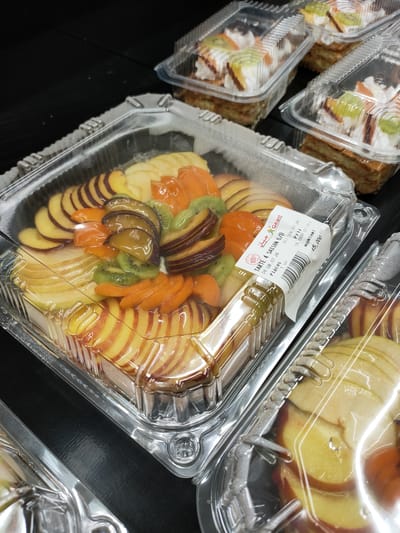 A close-up, high-angle shot shows a clear plastic container filled with sliced fruit, primarily peaches and possibly apples, along with slices of kiwi and apricot. The fruit is arranged in a circular pattern on top of a pastry base, covered in a clear glaze. A label on the side of the container reads "Tarte 4 Saison G/B" and "DULC.09 07 26", indicating it is a seasonal tart with a date of July 26th. Other nearby containers also hold pastries, some topped with whipped cream and fruit. The containers are placed on a dark, reflective surface. The lighting suggests an indoor environment, possibly a bakery or supermarket display. The image was taken in Cebalat, Tunisia, as indicated by the context.
