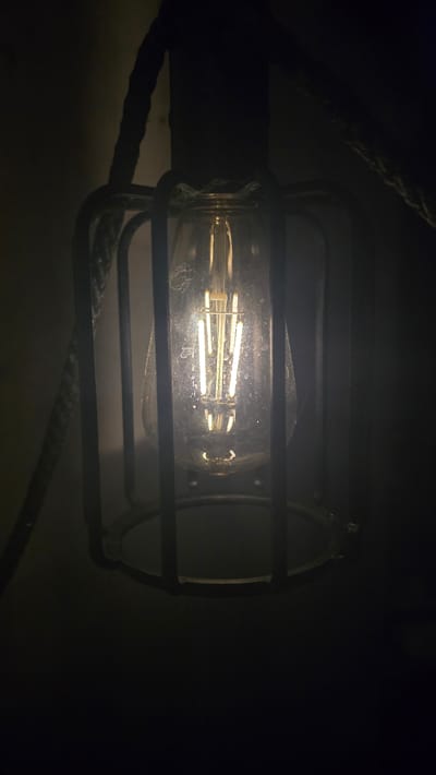 An Edison-style light bulb with a decorative cage is illuminated in a dimly lit environment. The light bulb has a clear glass casing and an LED filament that emits a warm yellow light. The cage is made of dark metal bars, enclosing the bulb. A thick rope, possibly part of the hanging mechanism, is visible in the upper left and right portions of the image. The background is dark and out of focus, suggesting a low-light setting. There is no visible indication of the specific location within the city of Garwolin, Poland.